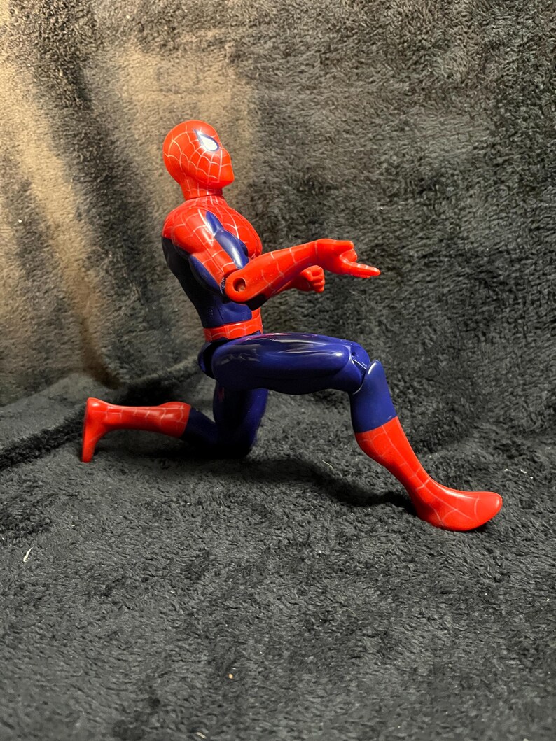 May include: A red and blue Spider-Man action figure in a crouching pose, pointing with one hand. The figure has a red head and feet, with a blue body and arms. The figure is on a dark surface.