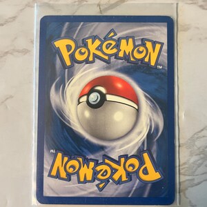 以下が含まれることがあります： 保護スリーブに入ったポケモンカード。カードの青い背景には、黄色のポケモンロゴと赤と白のモンスターボールのデザインがあります。カードは長方形です。