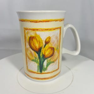 May include: Cream-colored ceramic mug with a white handle and rim. The mug features a rectangular design with yellow and orange floral artwork. The artwork includes yellow flowers with green stems and leaves, set against a light background.