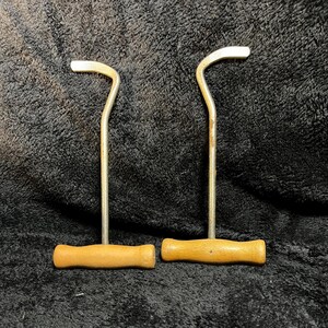May include: Two vintage metal tools with wooden handles. Each tool has a curved metal hook at the top and a cylindrical wooden handle at the bottom. The metal has a silver finish, and the wood is a light brown color.
