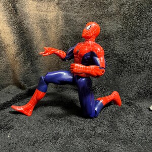 May include: A Spiderman action figure in a kneeling pose. The figure is primarily red and blue, with a detailed web pattern on the red sections. The figure is on a dark background.