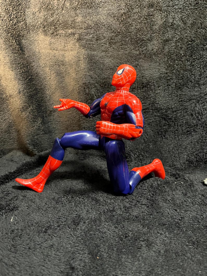 May include: A Spiderman action figure in a kneeling pose. The figure is primarily red and blue, with a detailed web pattern on the red sections. The figure is on a dark background.