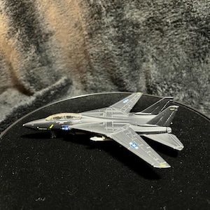 May include: A dark gray toy fighter jet with detailed wings, tail fins, and cockpit. The model has blue and white star insignias and yellow accents. The jet is displayed on a black surface, showcasing its design.