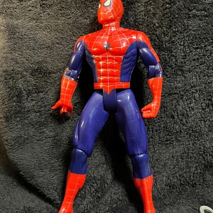 May include: A red, blue, and white Spider-Man action figure. The figure is in a standing pose with arms outstretched. The figure's head is red with white eye details, and the torso is red with a web pattern. The legs are blue and red.