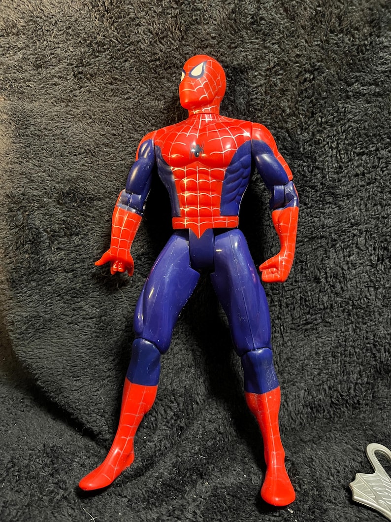 May include: A red, blue, and white Spider-Man action figure. The figure is in a standing pose with arms outstretched. The figure's head is red with white eye details, and the torso is red with a web pattern. The legs are blue and red.