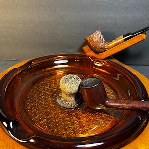 May include: An amber-colored glass ashtray with a textured interior, holding a brown pipe, a pipe stand, and a pipe tamper. The ashtray sits on a wooden surface, with a second pipe resting on a wooden stand.