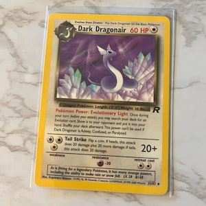 May include: A Dark Dragonair Pokémon card in a protective sleeve. The card features a purple and white dragon-like creature with a crystal background. The card text includes "Dark Dragonair 60 HP" and "Tail Strike". The card is 13' 1" long and weighs 36 lbs.
