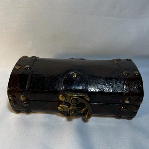 May include: A small, dark brown wooden chest with a curved top and brass-colored hardware. The chest is secured with a clasp and is adorned with small, round studs. The overall design suggests a vintage or antique style.