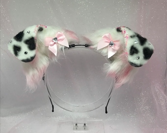 Könnte beinhalten: Ein weiß-schwarz geflecktes Stirnband mit rosa Fell und rosa Schleifen mit Strasssteinen. Das Stirnband liegt auf einer weißen Oberfläche.