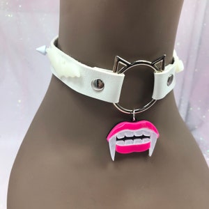 May include: White leather choker with silver cat ears and a silver ring. The choker has a pink and white vampire teeth charm.