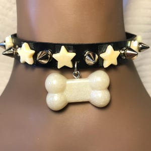 May include: Black leather dog collar with cream-colored star and spike accents. The collar features a bone-shaped charm. The stars and bone charm have a pearlescent finish. Silver-colored metal studs and spikes are also present.