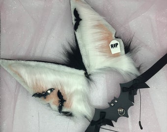 May include: White and pink faux fur cat ears with black bat accents and a black bat headband with a silver ring. The headband has a black bow on each side. The ears have a small white tag that says "RIP".