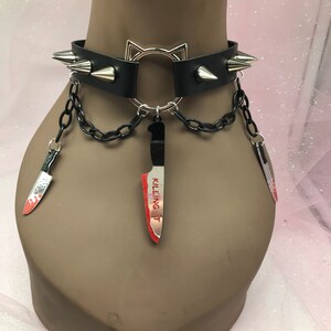 May include: Black leather choker with silver spikes and a cat-shaped ring. The choker has a chain with three dangling knife charms, one of which says "Killing It".