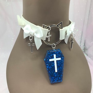 May include: White faux leather choker with a silver cat charm, a silver ring with a cross charm, and two white bows with silver cross charms. The choker also features a blue glitter coffin charm with a white cross.