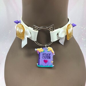 May include: White leather choker with silver cat ears and spikes. The choker has a charm attached to it that says "Stay Cute" with a purple tombstone and a pink heart.
