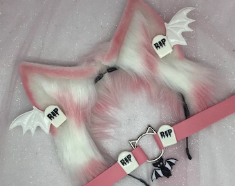 Pode incluir: Faixa de cabeça de orelhas de gato de pele sintética rosa e branca com asas de morcego brancas e pretas e um colar rosa com um anel prateado e um amuleto de morcego preto. O colar tem três pingentes de lápide branca com a palavra "RIP" neles.