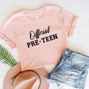 Puede incluir: Una camiseta rosa con el texto "Official Pre-Teen" en letras negras. La camiseta está colocada sobre una superficie blanca con unos pantalones cortos de mezclilla y un sombrero marrón.