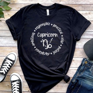 May include: Black t-shirt with a white circle design featuring the Capricorn zodiac symbol and the words "Capricorn", "responsible", "practical", "wise", "patient", "ambitious", "disciplined", and "determined".