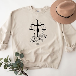 May include: A beige sweatshirt with a black Libra zodiac symbol, floral design, and the word "LIBRA" printed on the front.