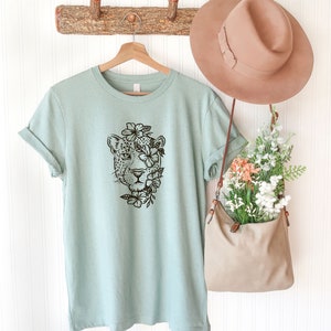 May include: A light blue t-shirt with a black and white graphic of a leopard's face surrounded by flowers. The shirt is hanging on a wooden hanger against a white wall.