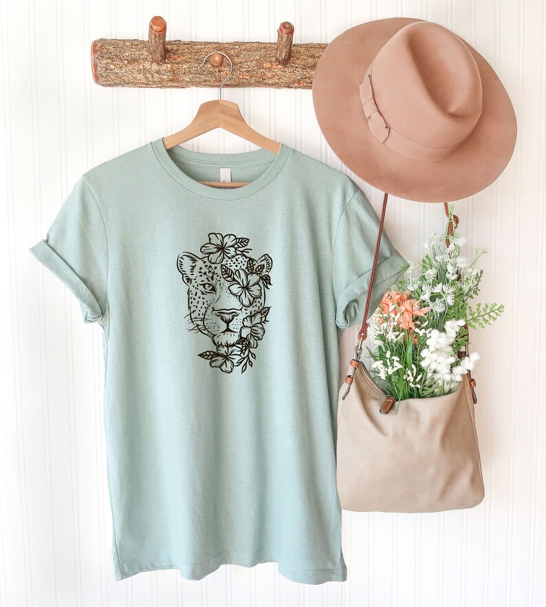 May include: A light blue t-shirt with a black and white graphic of a leopard's face surrounded by flowers. The shirt is hanging on a wooden hanger against a white wall.