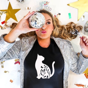 Może przedstawiać: Kobieta w czarnym t-shircie z białym sylwetką kota z kwiatowymi detalami. Trzyma dyskotekowy globus przed twarzą i jest otoczona konfetti i innymi dekoracjami imprezowymi. Na koszulce widnieje napis "Happy New Year".
