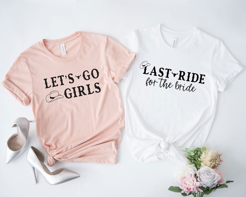 Op de afbeelding: Twee roze en witte t-shirts met zwarte tekst. Het roze shirt zegt "Let's Go Girls" met een cowboyhoed-afbeelding. Het witte shirt zegt "Last Ride for the Bride" met een cowboyhoed-afbeelding.