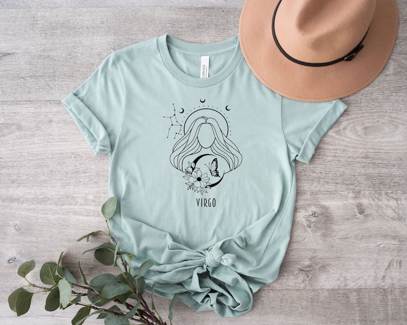 Puede incluir: Una camiseta azul claro con un dise&ntilde;o gr&aacute;fico en blanco y negro de una mujer con una mariposa y flores. El gr&aacute;fico tambi&eacute;n incluye el texto "VIRGO".