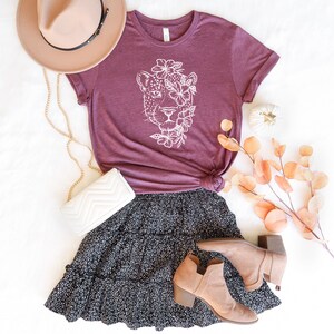 May include: A maroon t-shirt with a white graphic of a leopard's face surrounded by flowers. The shirt is paired with a black and white polka dot skirt and brown ankle boots.