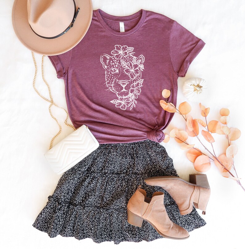 May include: A maroon t-shirt with a white graphic of a leopard's face surrounded by flowers. The shirt is paired with a black and white polka dot skirt and brown ankle boots.