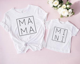 May include: Two white t-shirts with black text. The larger shirt says "MAMA" in a square box. The smaller shirt says "MINI" in a square box.
