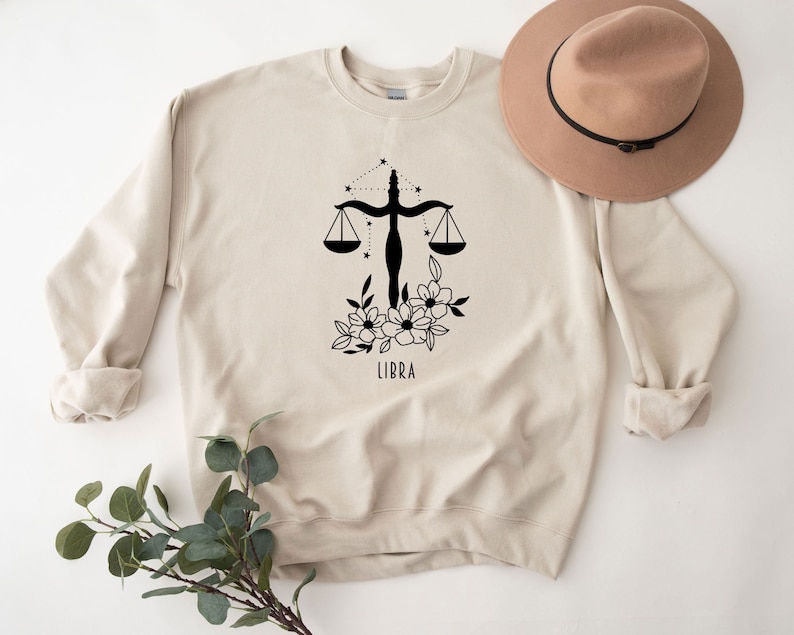 May include: A beige sweatshirt with a black Libra zodiac symbol, floral design, and the word "LIBRA" printed on the front.