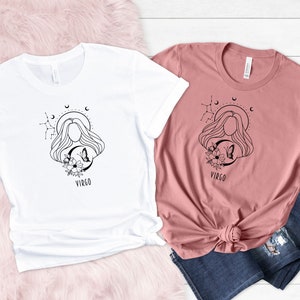 Puede incluir: Dos camisetas con un dise&ntilde;o del signo del zod&iacute;aco Virgo. El dise&ntilde;o presenta a una mujer con el pelo suelto, una luna creciente, estrellas y flores. La palabra "Virgo" est&aacute; impresa debajo del dise&ntilde;o. Una camiseta es blanca y la otra es de color rosa polvoriento.