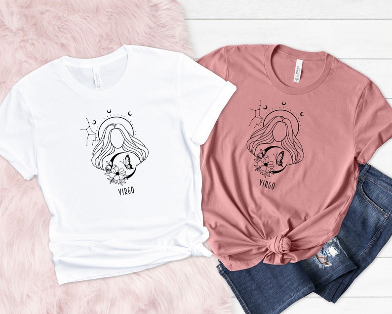 Puede incluir: Dos camisetas con un dise&ntilde;o del signo del zod&iacute;aco Virgo. El dise&ntilde;o presenta a una mujer con el pelo suelto, una luna creciente, estrellas y flores. La palabra "Virgo" est&aacute; impresa debajo del dise&ntilde;o. Una camiseta es blanca y la otra es de color rosa polvoriento.