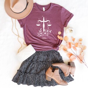 May include: A maroon t-shirt with a white floral Libra zodiac sign graphic. The shirt is paired with a black and white polka dot skirt, a white purse, a brown hat, and brown ankle boots.