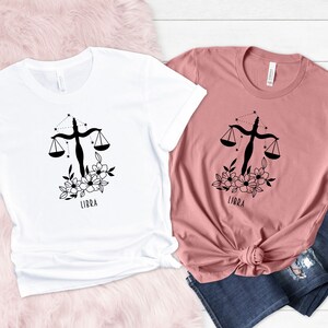 May include: Two t-shirts with a black and white floral design featuring the Libra zodiac symbol and the word "Libra". One t-shirt is white and the other is pink.