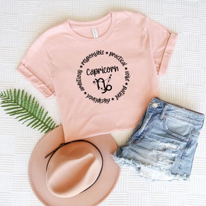 May include: A pink t-shirt with a black circle design featuring the zodiac symbol for Capricorn and the words "Capricorn", "ambitious", "disciplined", "responsible", "practical", "patient", "wise".