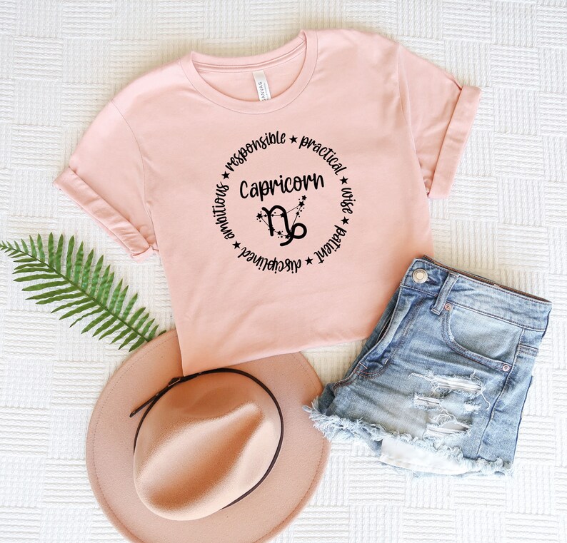 May include: A pink t-shirt with a black circle design featuring the zodiac symbol for Capricorn and the words "Capricorn", "ambitious", "disciplined", "responsible", "practical", "patient", "wise".