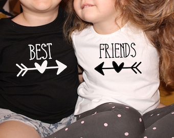 Puede incluir: Dos niños con camisas a juego con flechas negras y corazones blancos. La camisa de la izquierda dice "BEST" y la camisa de la derecha dice "FRIENDS".