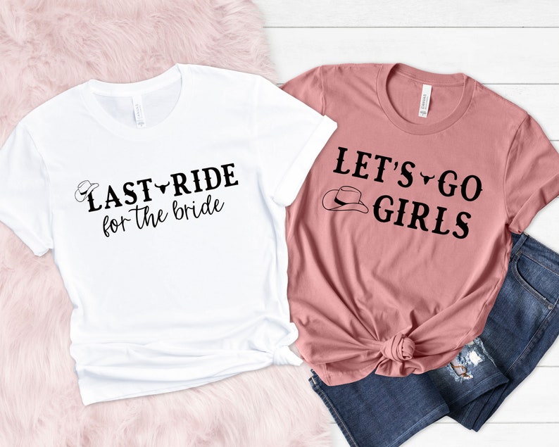 Op de afbeelding: Twee T-shirts met een western thema. Het witte T-shirt zegt "Last Ride for the Bride" met een cowboyhoed en een stierenkop-afbeelding. Het roze T-shirt zegt "Let's Go Girls" met een cowboyhoed-afbeelding.