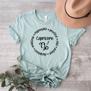 May include: A light blue t-shirt with a black circle graphic featuring the Capricorn zodiac symbol and the words "Capricorn", "ambitious", "responsible", "practical", "wise", "patient", "achiever".