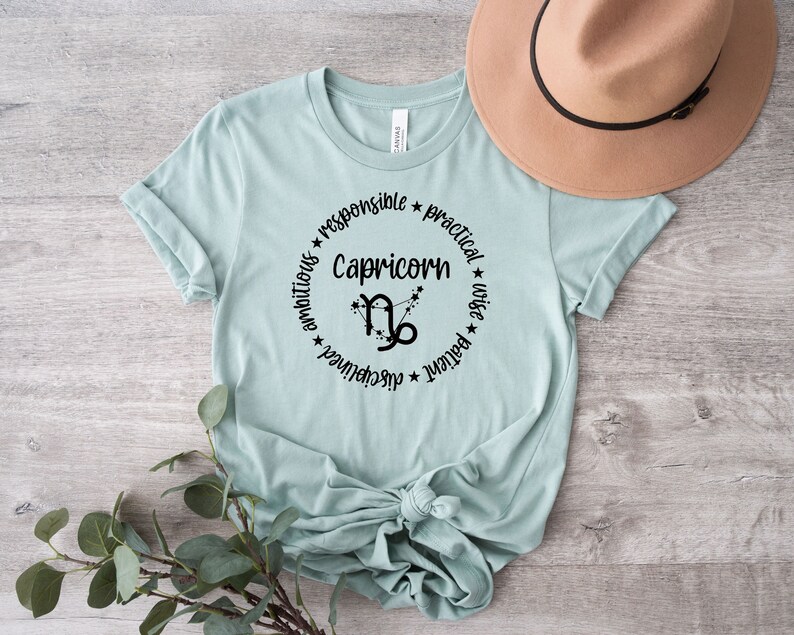 May include: A light blue t-shirt with a black circle graphic featuring the Capricorn zodiac symbol and the words "Capricorn", "ambitious", "responsible", "practical", "wise", "patient", "achiever".