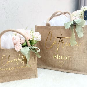 May include: Two burlap tote bags with gold lettering. One bag says "Charlotte Maid of Honour" and the other says "Catherine Bride".