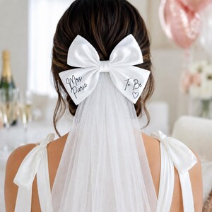 May include: A white satin hair bow with a sheer tulle veil attached. The left side of the bow features the black text "Mrs Pierce" and the right side features the text "To Be" with a small heart symbol. The bow is secured in dark brown hair.