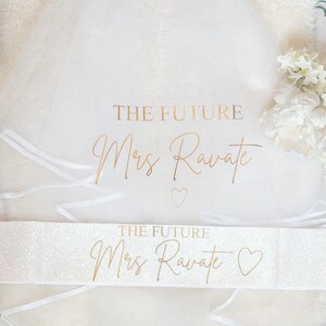 May include: A white tulle veil and a white glittery sash. The veil features the gold text THE FUTURE Mrs Ravate and a small heart. The sash also displays the gold text THE FUTURE Mrs Ravate with a heart symbol. Both items are white.