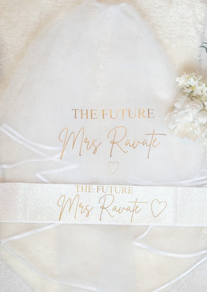 May include: A white tulle veil and a white glittery sash. The veil features the gold text THE FUTURE Mrs Ravate and a small heart. The sash also displays the gold text THE FUTURE Mrs Ravate with a heart symbol. Both items are white.