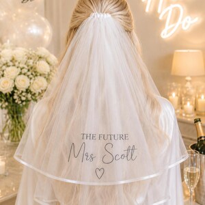 May include: A white two-tier bridal veil featuring a satin ribbon edge and the black printed text THE FUTURE Mrs Scott with a small heart symbol below. The veil is gathered at the top and worn over long blonde hair, draping down the back.