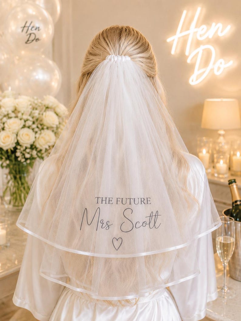 May include: A white two-tier bridal veil featuring a satin ribbon edge and the black printed text THE FUTURE Mrs Scott with a small heart symbol below. The veil is gathered at the top and worn over long blonde hair, draping down the back.