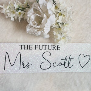 May include: A white glittery sash featuring the black printed text THE FUTURE Mrs Scott followed by a heart symbol. The sash has a shimmering, reflective surface and is displayed horizontally with white floral accents positioned above the text.