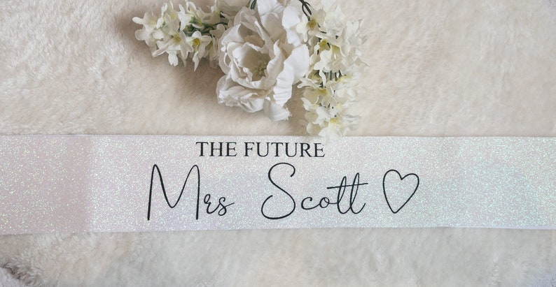 May include: A white glittery sash featuring the black printed text THE FUTURE Mrs Scott followed by a heart symbol. The sash has a shimmering, reflective surface and is displayed horizontally with white floral accents positioned above the text.